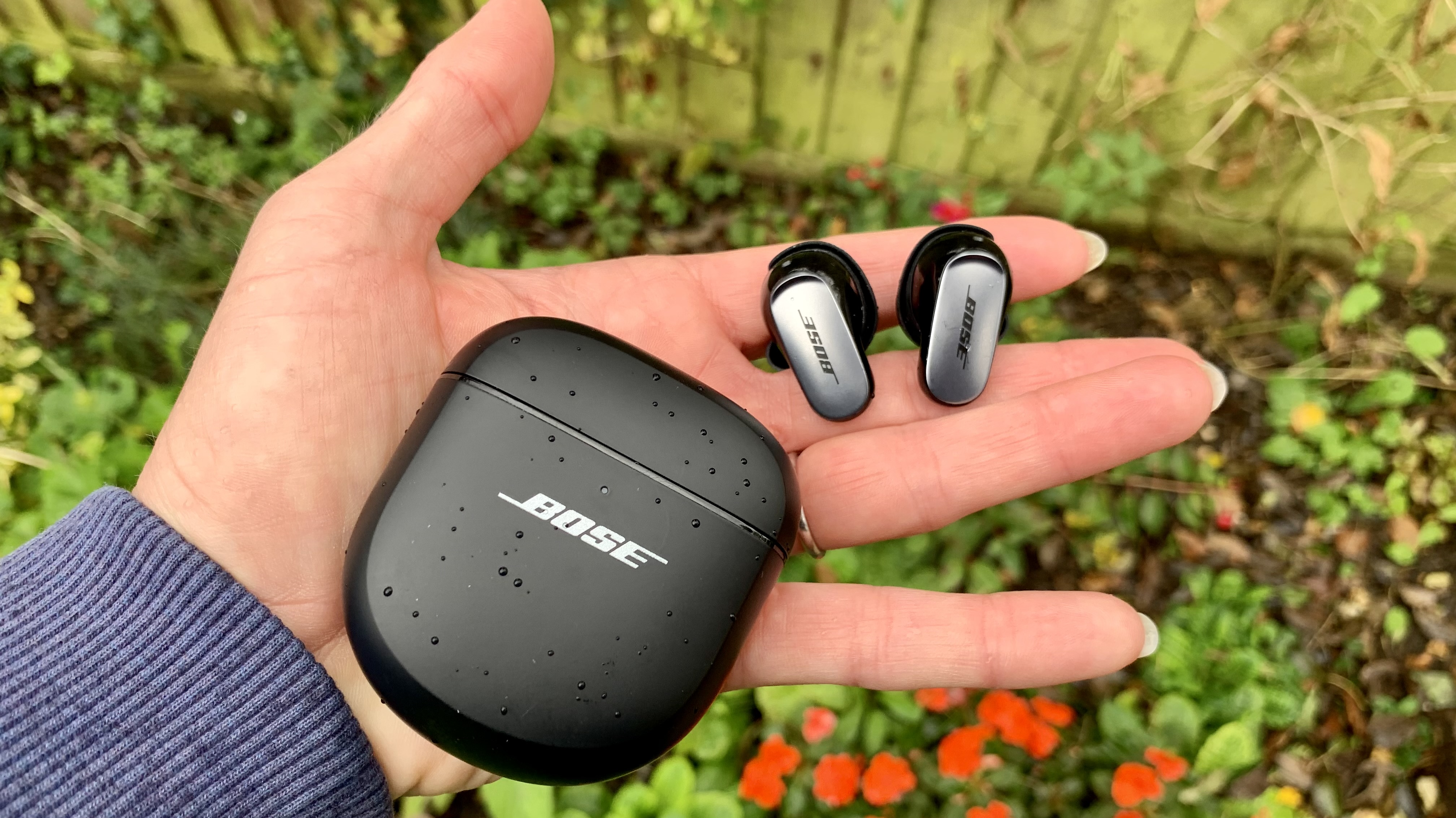 Bose QuietComfort Ultra earphones held in hand with case over a flower bed