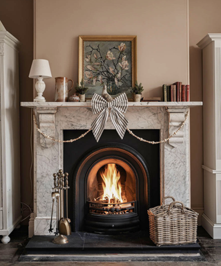 A white fireplace with a fire lit in it and a white lamp, floral painting, books on top of it, with a putty colored wall behind it