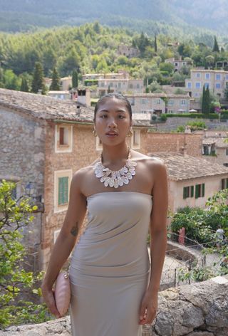 Shell jewelry trend shown on a woman wearing a white shell necklace with a strapless tube dress, a clutch bag, and hoop earrings.