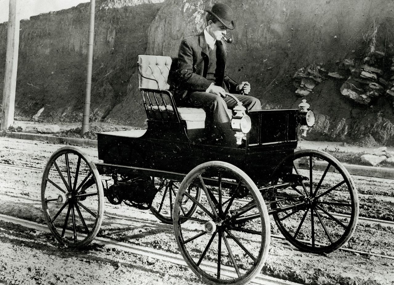 a black and white photo of an early 20th-century man driving a simple electric car