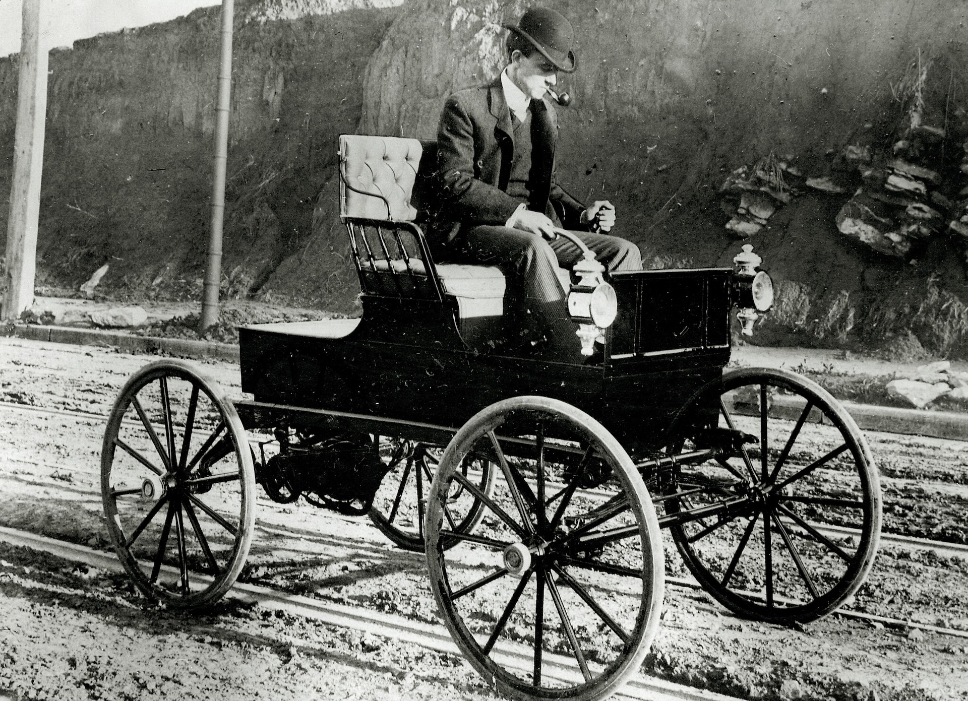 a black and white photo of an early 20th-century man driving a simple electric car