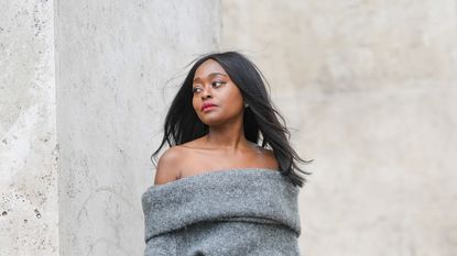 Carrole Sagba wears a gray oversized off shoulder pullover, pale gray suit pants with white feathers on one half leg, black and beige striped leather heels ankle boots, a white matte leather handbag, on April 18, 2021 in Paris, France.