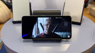 The Samsung Galaxy S25 in landscape orientation on a stand on a display table. On screen is Ryan Reynolds as the character Deadpool.