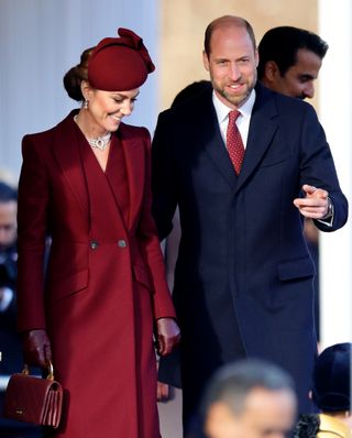 Kate Middleton wearing a maroon coat and hat standing next to Prince William in a navy coat and red tie and smiling