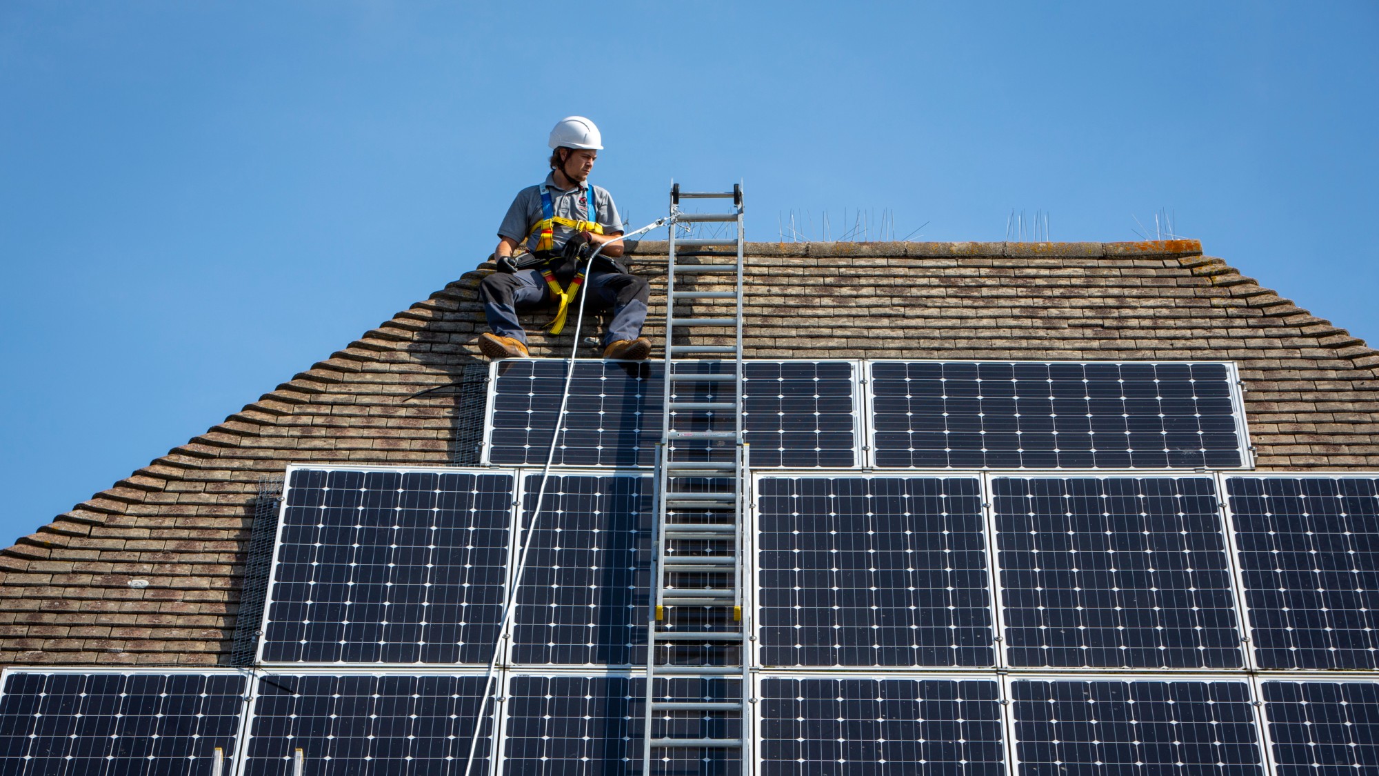 A maintenance man uses a ladder and harnesses to install equipment around a solar panel array on the roof of a house
