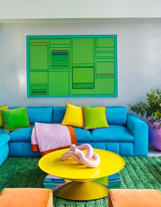 Grey living room with bright blue sofa, green rug and artwork, yellow coffee table and multicolor cushions