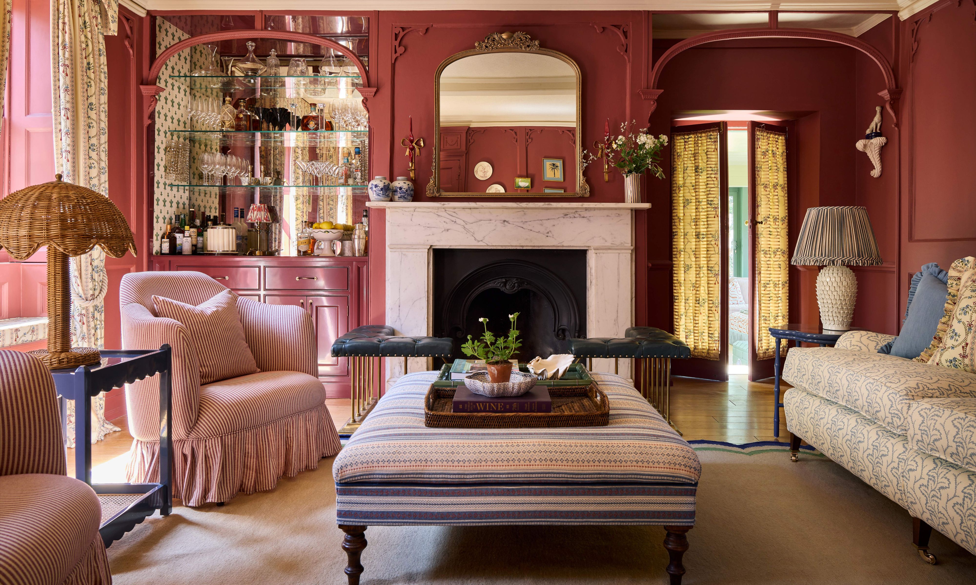 a red-pink painted historic drawing room with marble fireplace, custom home bar, paneled walls and layered furniture and patterns