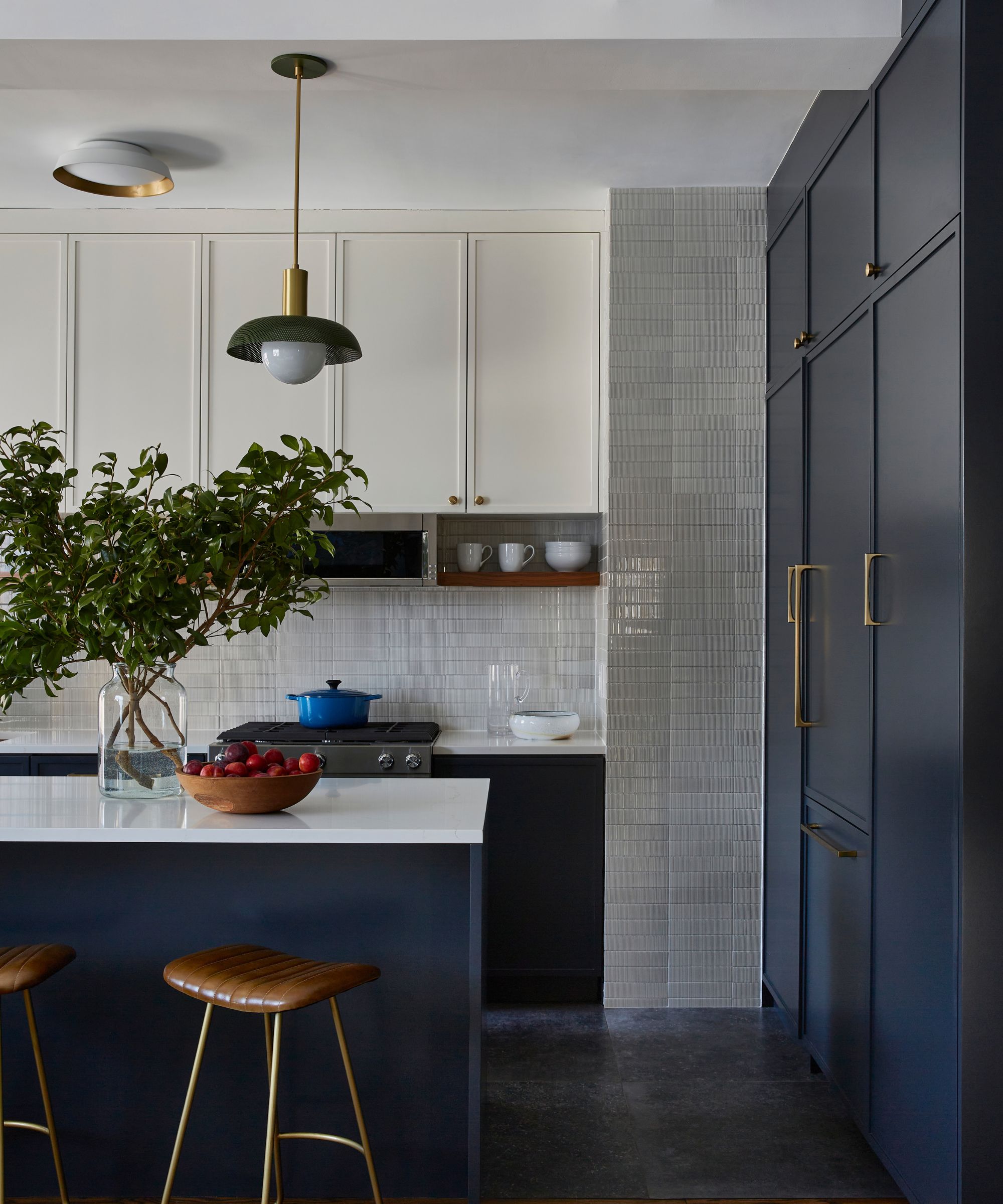 A modern kitchen with dark navy blue cabinets and an island. Gray wall tiles, and a row of white upper kitchen cabinets. Brass fixtures on the cabinets, and two tan leather bar stools by the island, and a vase of foliage.