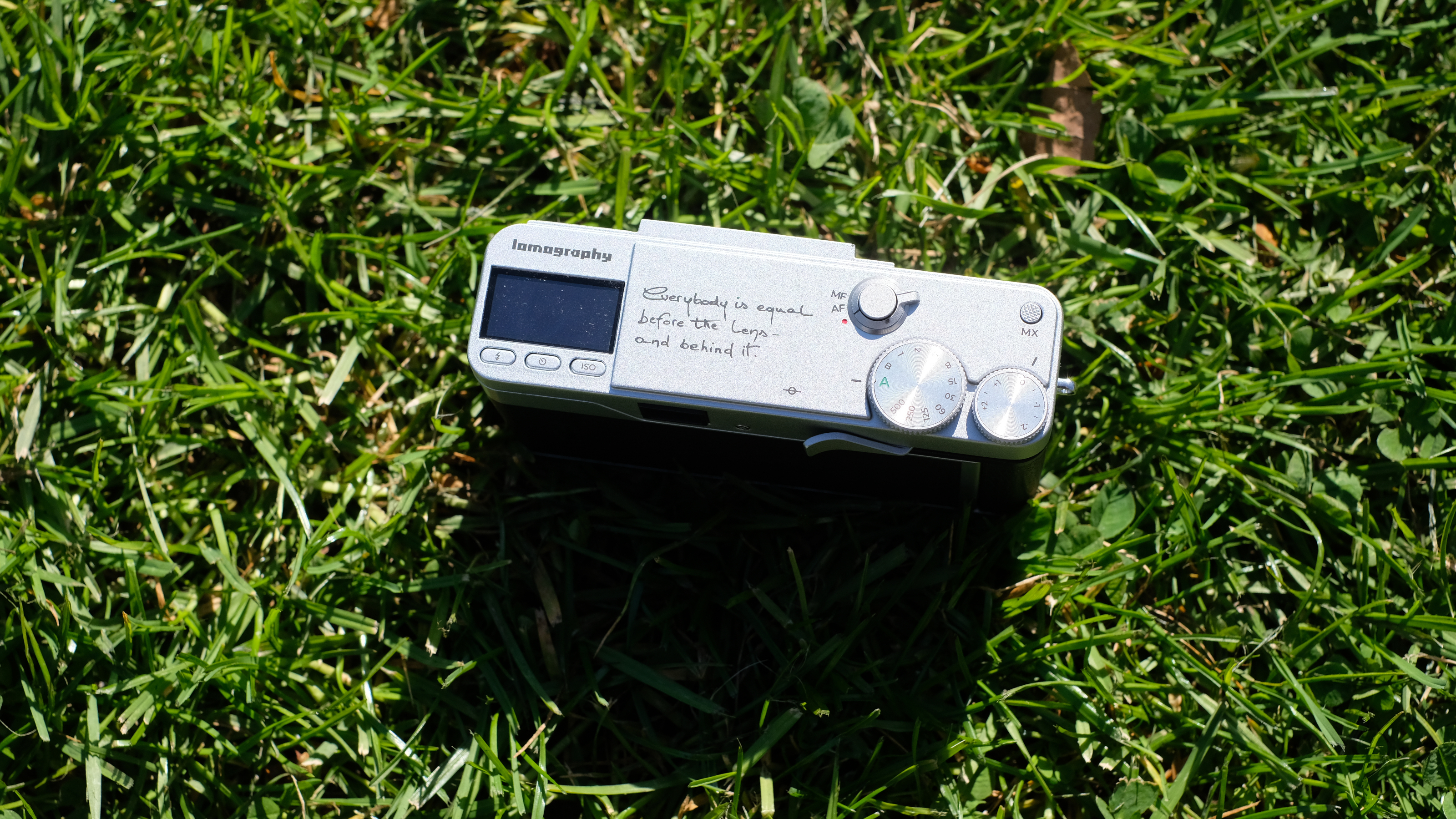 Photograph of the Lomography Lomo MC-A, with grass visible in background, showing wider view of top plate with engraving and dial controls
