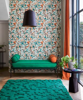 Green upholstered ottoman in front of abstract wallpapered wall, wooden floor and floor to ceiling windows.