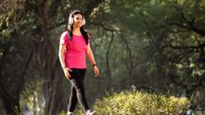 A woman walks outside wearing chunky over-ear headphones. Behind her we see green leafy trees.