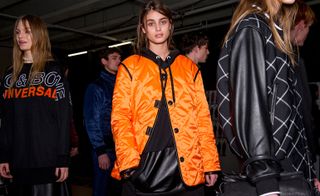 Main figure is a female model wearing a padded orange jacket over a black hoodie. Other models are in the background
