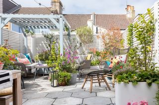 Modern garden ideas: a paved garden with light blue painted raised planters and pergola, hexagonal paving, a grey mesh chair and footstool and light grey slatted fence