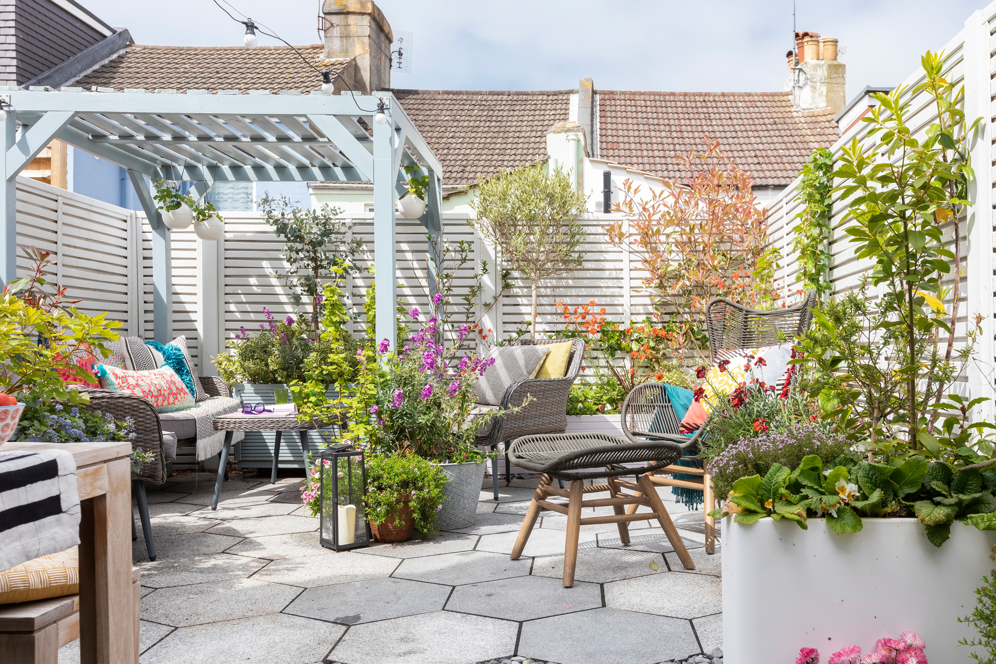 paved garden with light blue painted raised planters and pergola, hexagonal paving, a grey mesh chair and footstool and light grey slatted fence