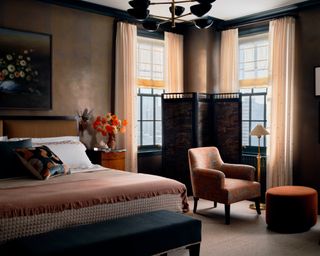 bedroom in earthy tones with coppery metallic walls, dark wood bed with upholstered headboard, dark end of bed bench, burnt orange chair and stool and screen