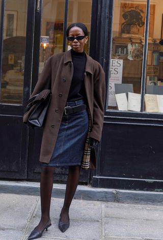 Black turtleneck outfit is shown in a photo of a French woman on the sidewalk wearing a brown coat over a black turtleneck styled with a denim skirt, black belt, black tights, black slingback heels, and a black bag