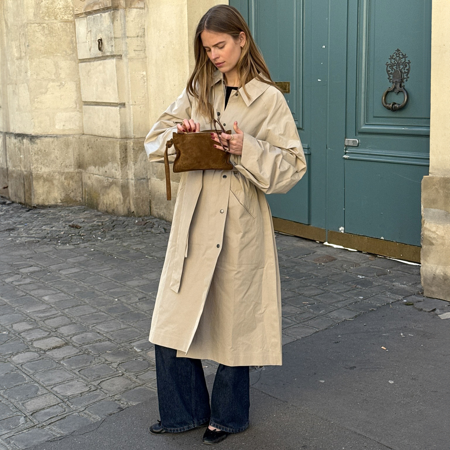 Scandi style influencer Clara Dyrhauge poses in Paris, France on a cobblestone street in front of an ornate teal green door wearing a calf-length tan trench coat, brown suede clutch bag, dark-wash flared jeans, and black ballet flats