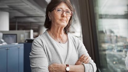 An older woman stands in an office with arms crossed.