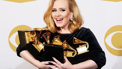 Adele smiling as she struggles to hold a collection of award trophies.
