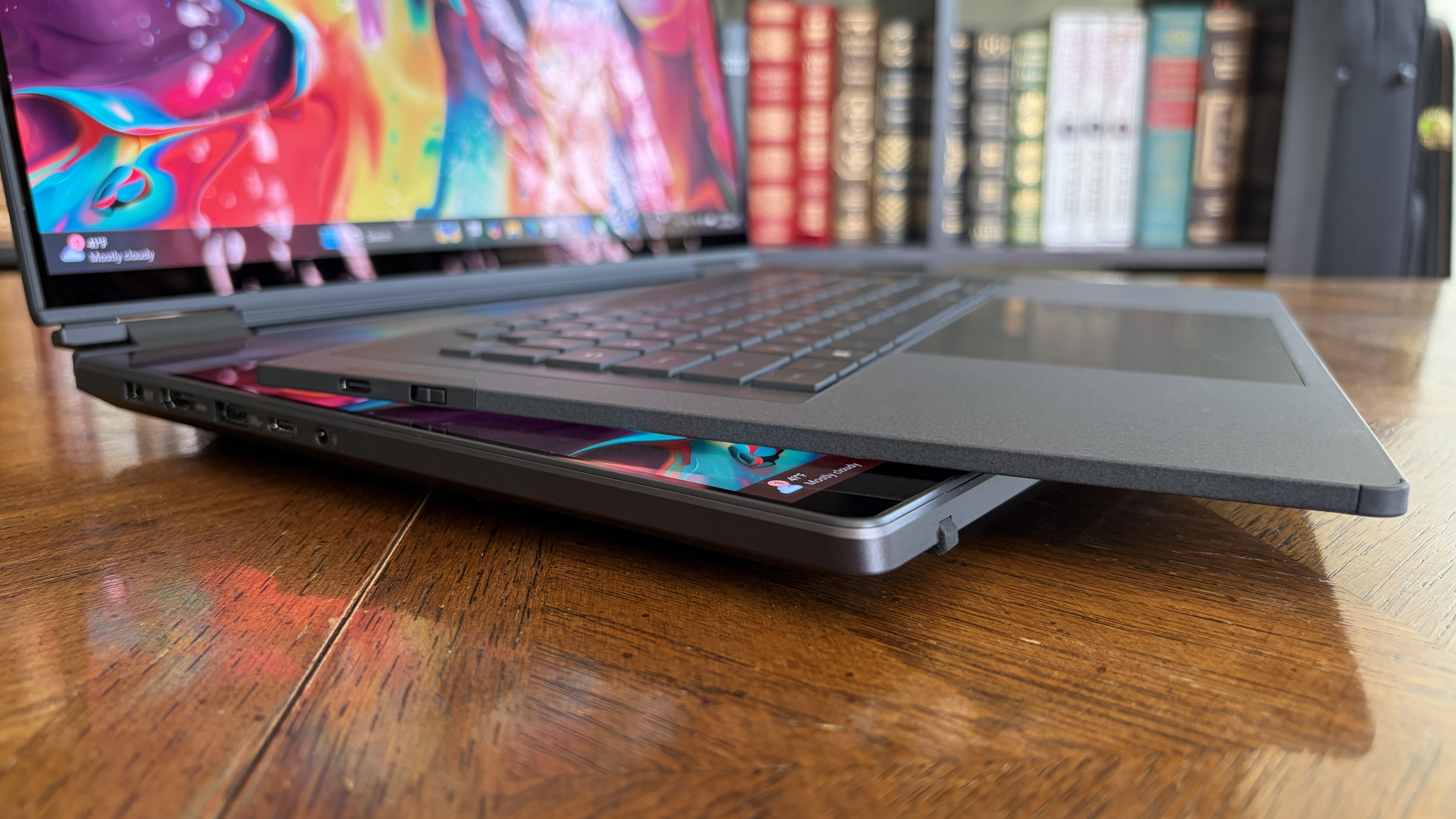 A sleek laptop with a vibrant screen partially open on a wooden table, with colorful books blurred in the background, conveying a modern and creative atmosphere.