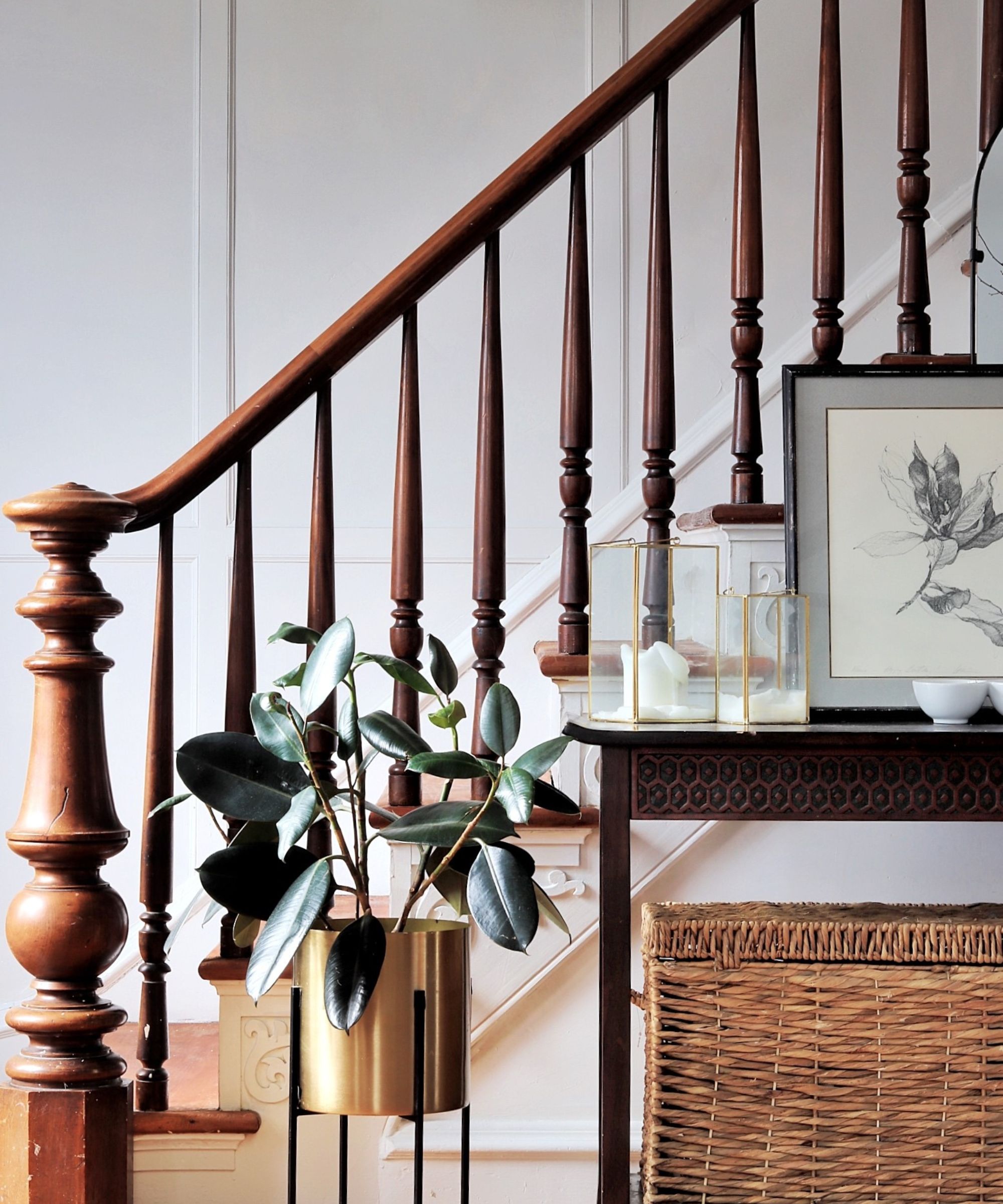 Side shot of staircase and wooden banister in background. Pictured in the foreground is a plant with a golden vase, a wicker storage box, and a side table with glass ornaments and a framed floral drawing.