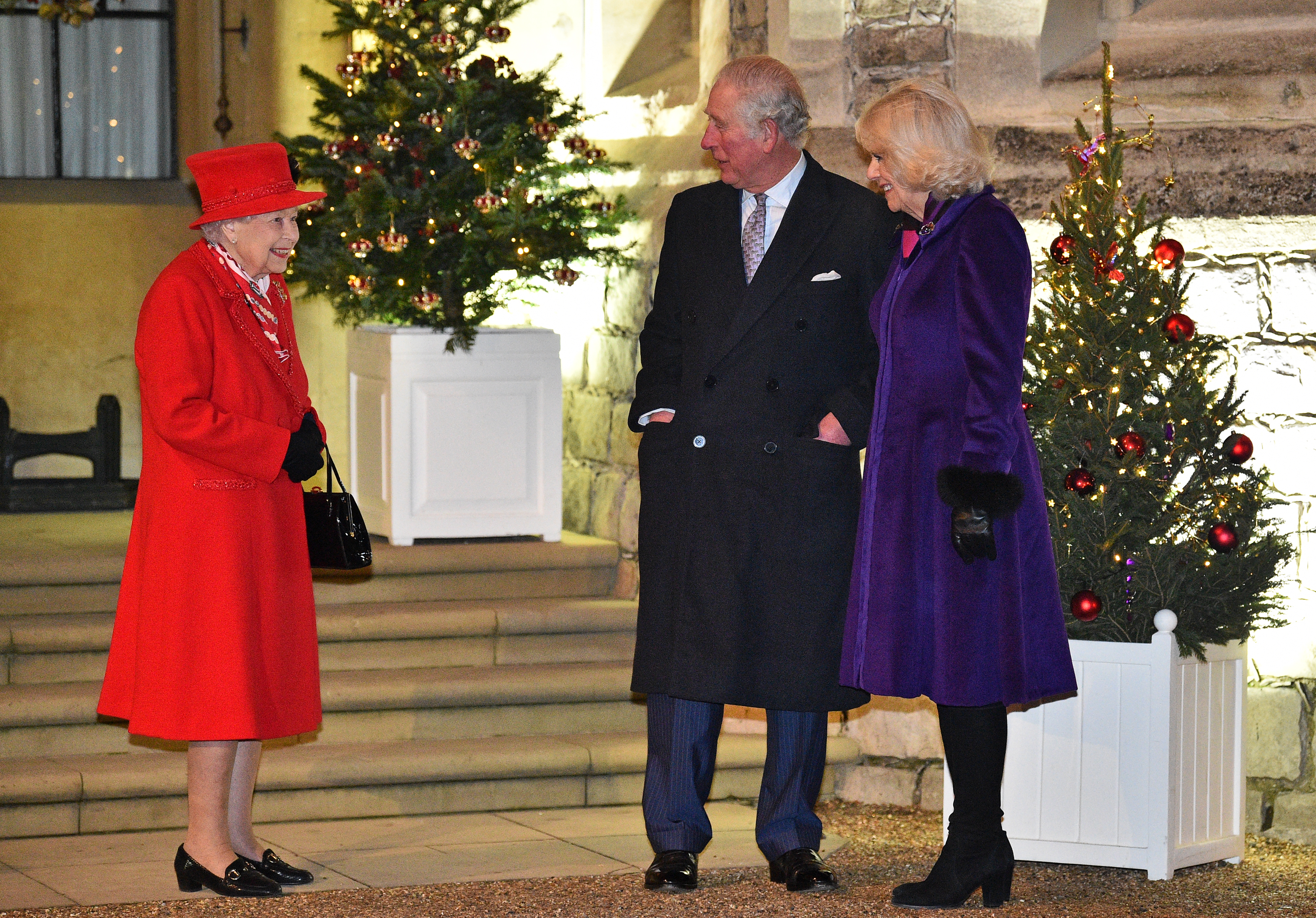 Queen Elizabeth wearing a red coat, smiling at King Charles and Queen Camilla in front of two Christmas trees at Windsor Castle in 2020