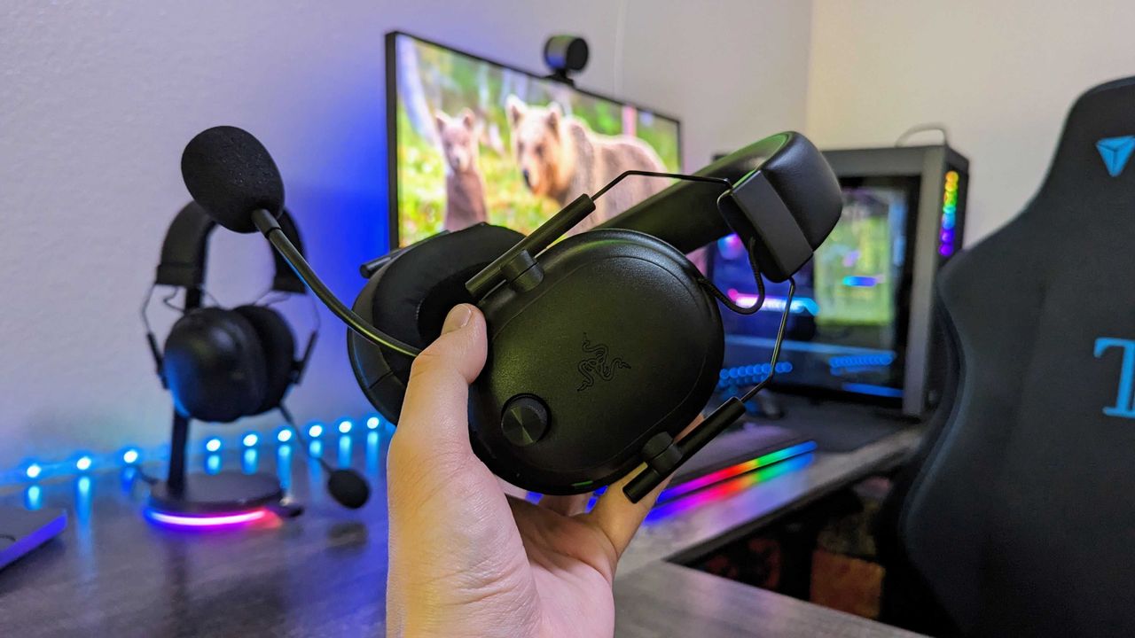 Hand holding black gaming headset with microphone in a setup featuring a colorful backlit keyboard, monitor displaying bear cubs, and gaming chair.