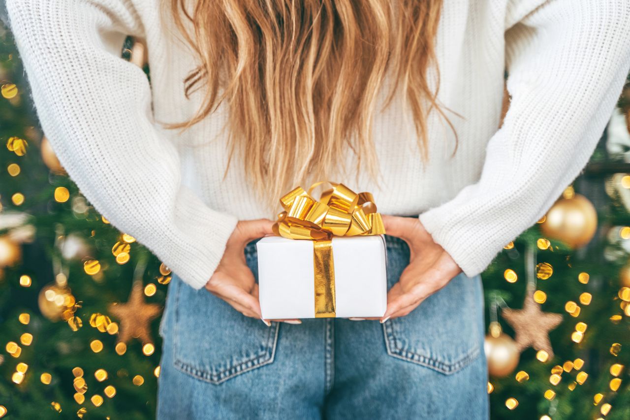 A woman holding a small white present with a gold bow behind her back in front of a decorated Christmas tree.