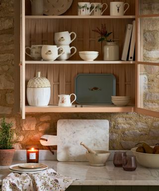 A kitchen pantry with a wooden shelf with cups in it and a kitchen counter with a marble top and a candle on it