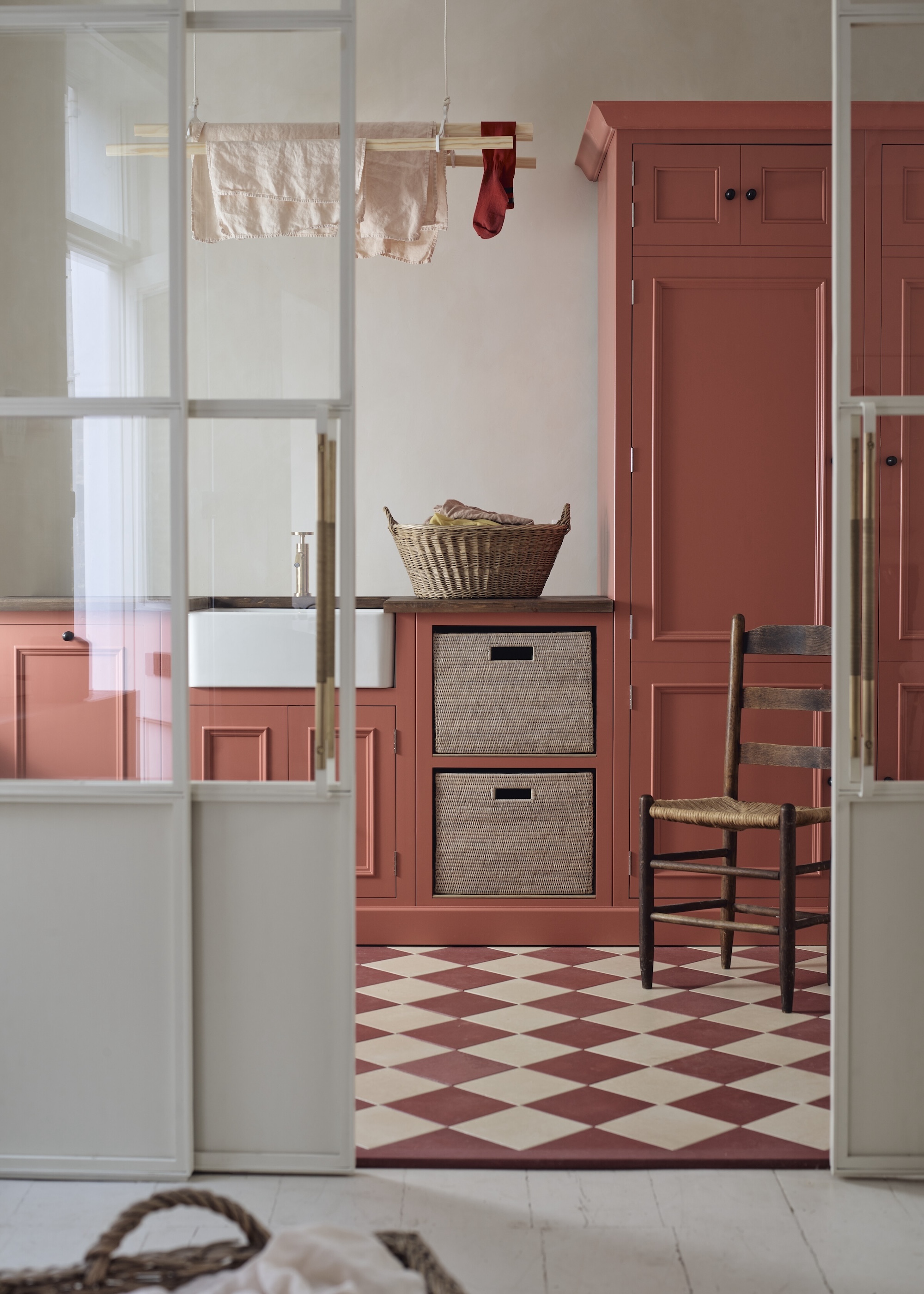 A warm and inviting coral colored utility room with tall cupboard storage, pull out baskets and a ceiling airer with classic chequerboard tiled flooring