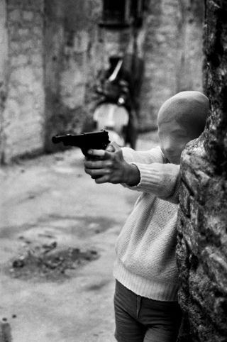 Black and white photograph of a young boy with a thin balaclava-like disguise over his head. He is aiming a handgun at something