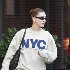 Hailey Bieber wearing an NYC sweatshirt, stovepipe jeans, a black The Row bag, wraparound sunglasses, and black leather flip-flops in Beverly Hills
