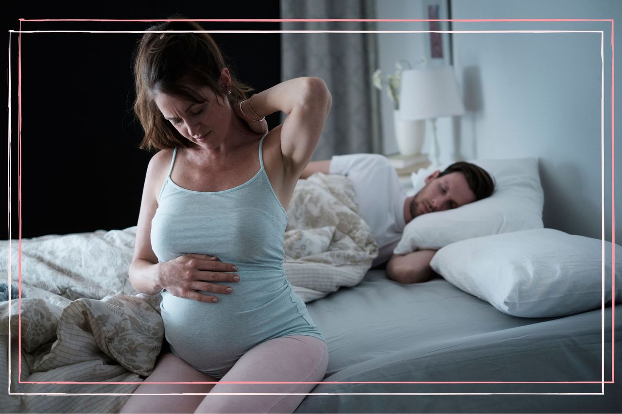 A pregnant woman sat up in bed while her partner sleep behind her