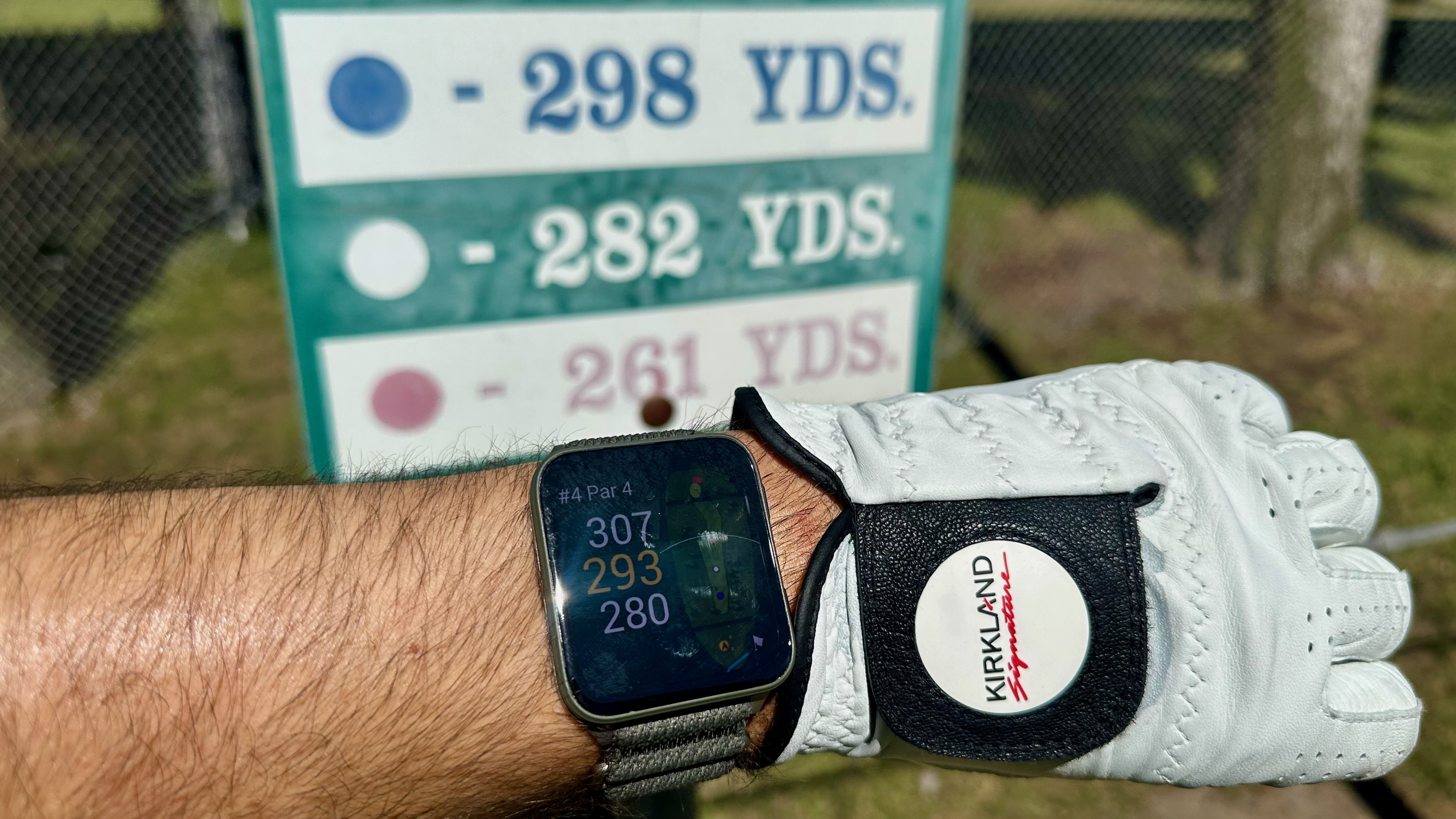 The Garmin Venu X1 on a wrist next to a golf glove, showing the yardage for the hole, a golf sign showing the actual yardage behind the Venu.