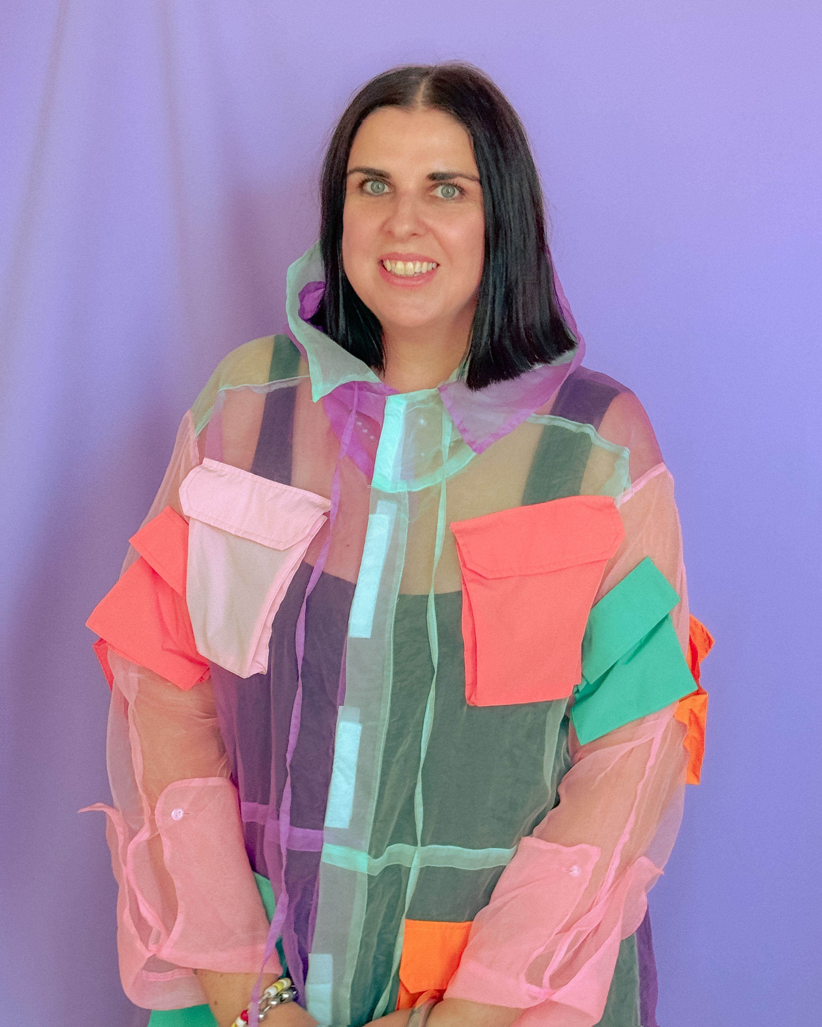 Two people dressed in colorful clothing stand against an electric lilac background while smiling to the camera.