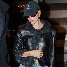 Hailey Bieber during Paris Fashion Week wearing a fitted, fringed leather jacket, straight-leg jeans, high-heel mules, a Mercedes-Benz baseball cap, and sunglasses