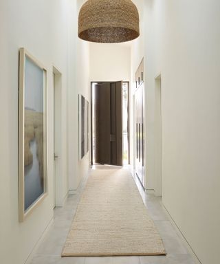 Calming hallway space with light gray stone flooring, white painted walls decorated with large framed artwork, skylight in ceiling, large textured, woven pendant, beige runner on floor, dark brown front door