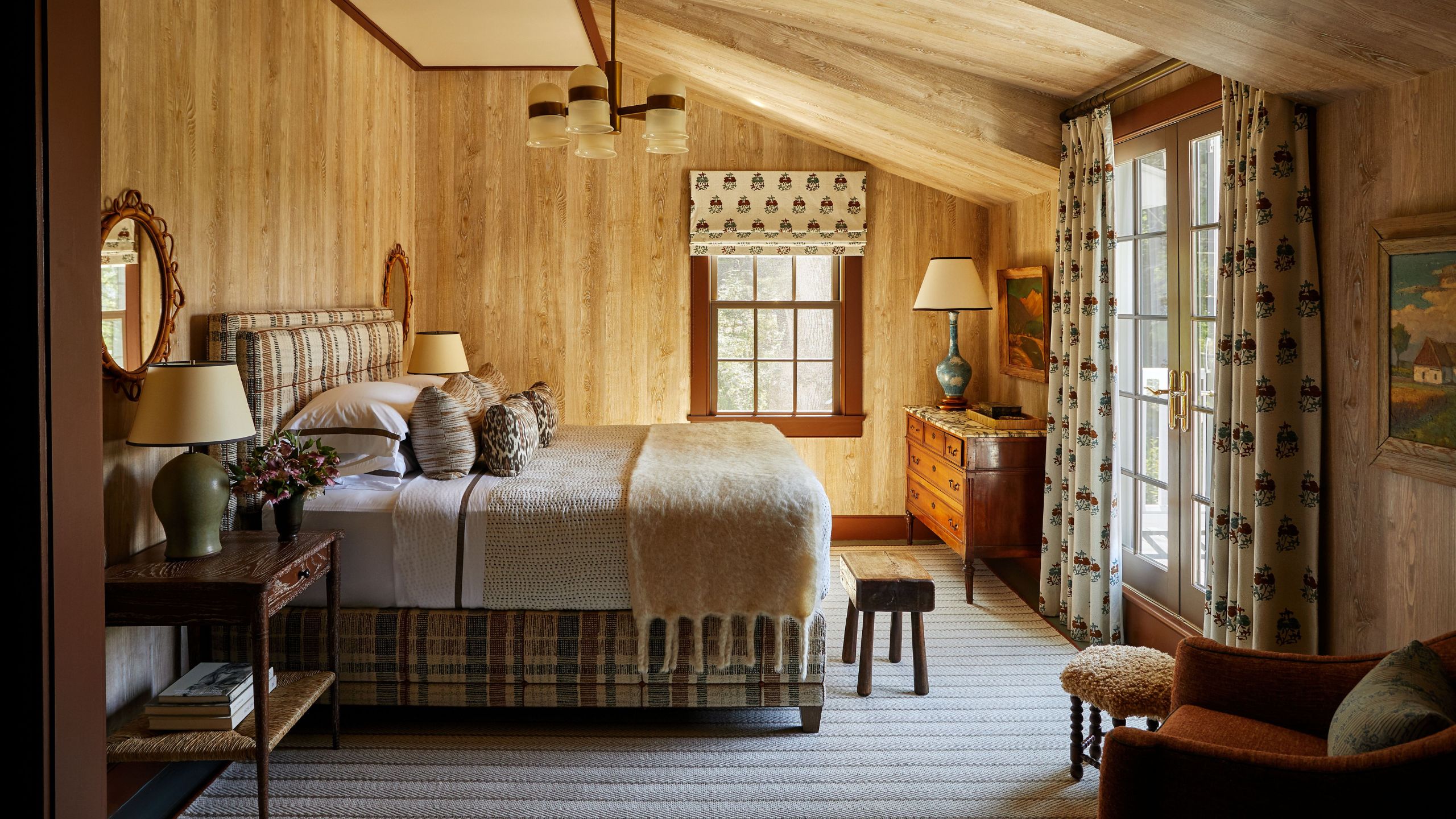 a timber panelled primary bedroom with large french doors framed by floral drapes, with a plaid upholstered bed in the center styled with natural textured linens