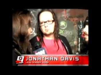 Long-haired man with glasses engages in conversation, holding a microphone. A name caption at the bottom reads "Jonathan Davis, Lead Singer, Korn." The background is a promotional setting.