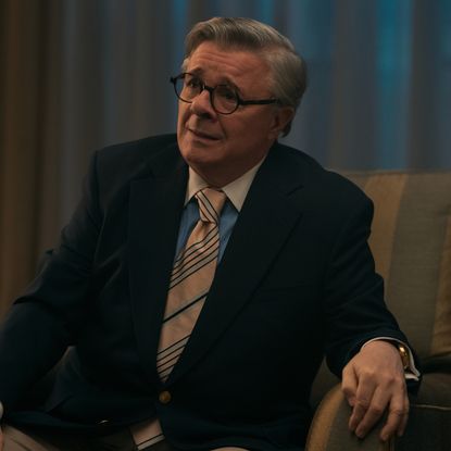 Nathan Lane as Dominick Dunne, sitting in an armchair with a glass on a table next to him, in episode 207 of Monsters: The Lyle And Erik Menendez Story.