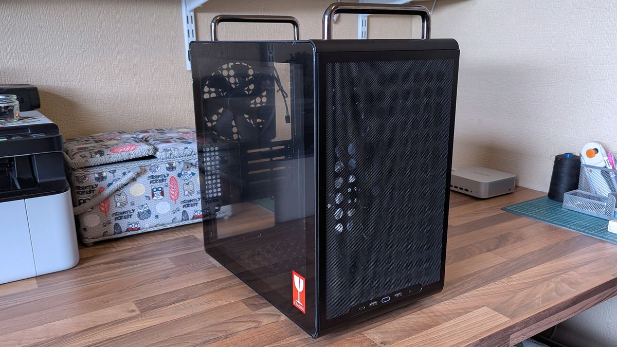 A Cooler Master Qube 340 case rests on a desk with its tempered glass side panel attached, revealing the empty interior and rear cooling fan behind the transparent panel.