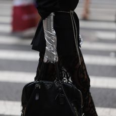 A Fashion Week guest is seen wearing big silver hoop earrings, black sunglasses from Celine, a long black blazer with a rhinestone ribbon as a belt, underneath a transparent skirt with black pattern, shiny silver gloves, a black leather bag from Prada and black leather heels from Maison Margiela outside before the Christian Siriano Show on September 08, 2023 in New York City.