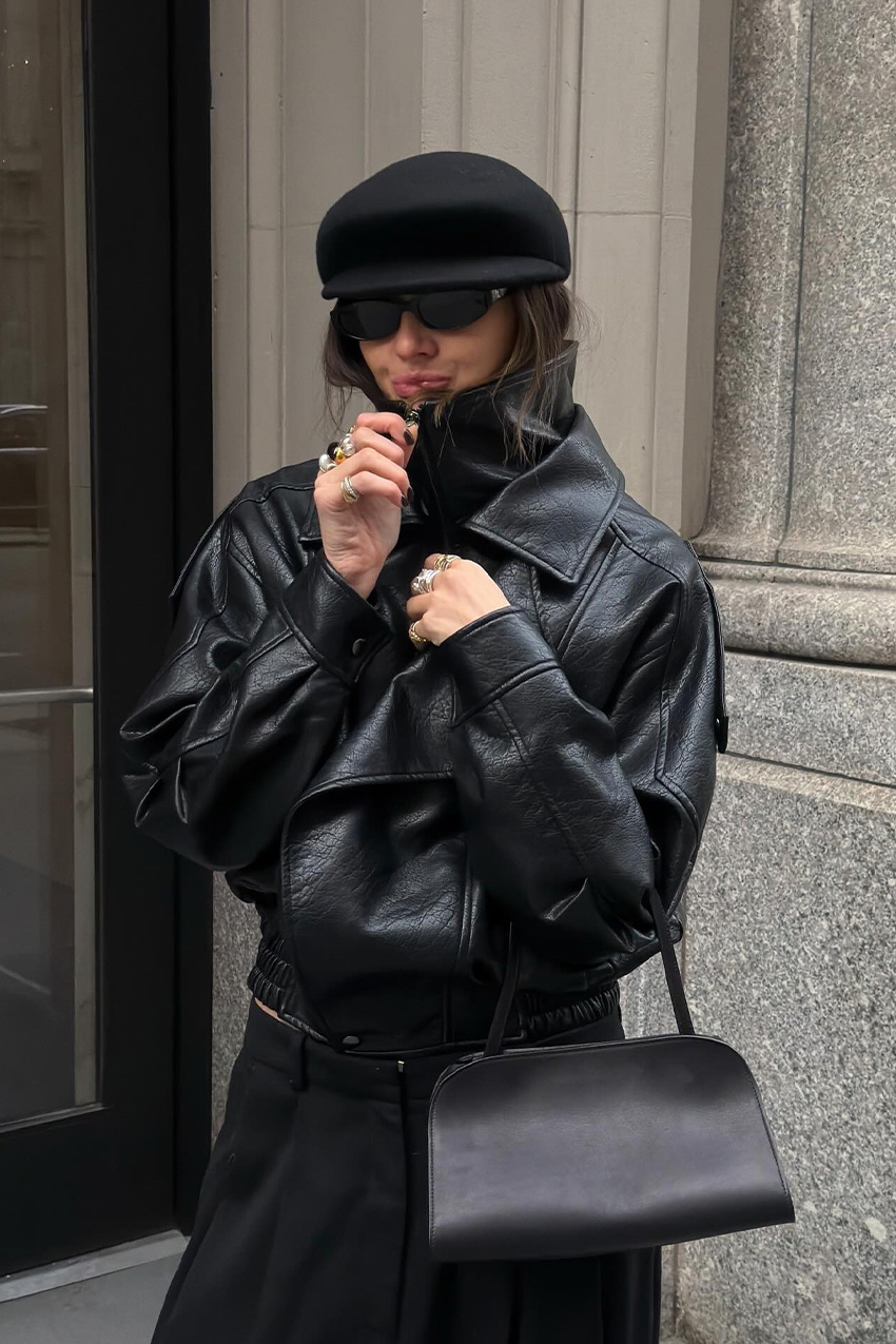 West Village winter trends are shown in a close street-style photo of a woman standing in front of a glass doorway in a concrete building in her winter outfit. She's wearing a black leather funnel neck bomber jacket with black trousers, black sunglasses, a black baker cap, layered silver rings, and a black bowler bag.