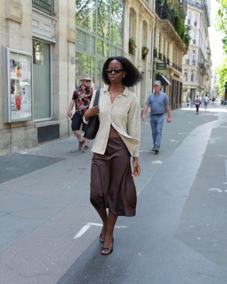 Woman on street wears beige shirt, brown slip skirt and black heels