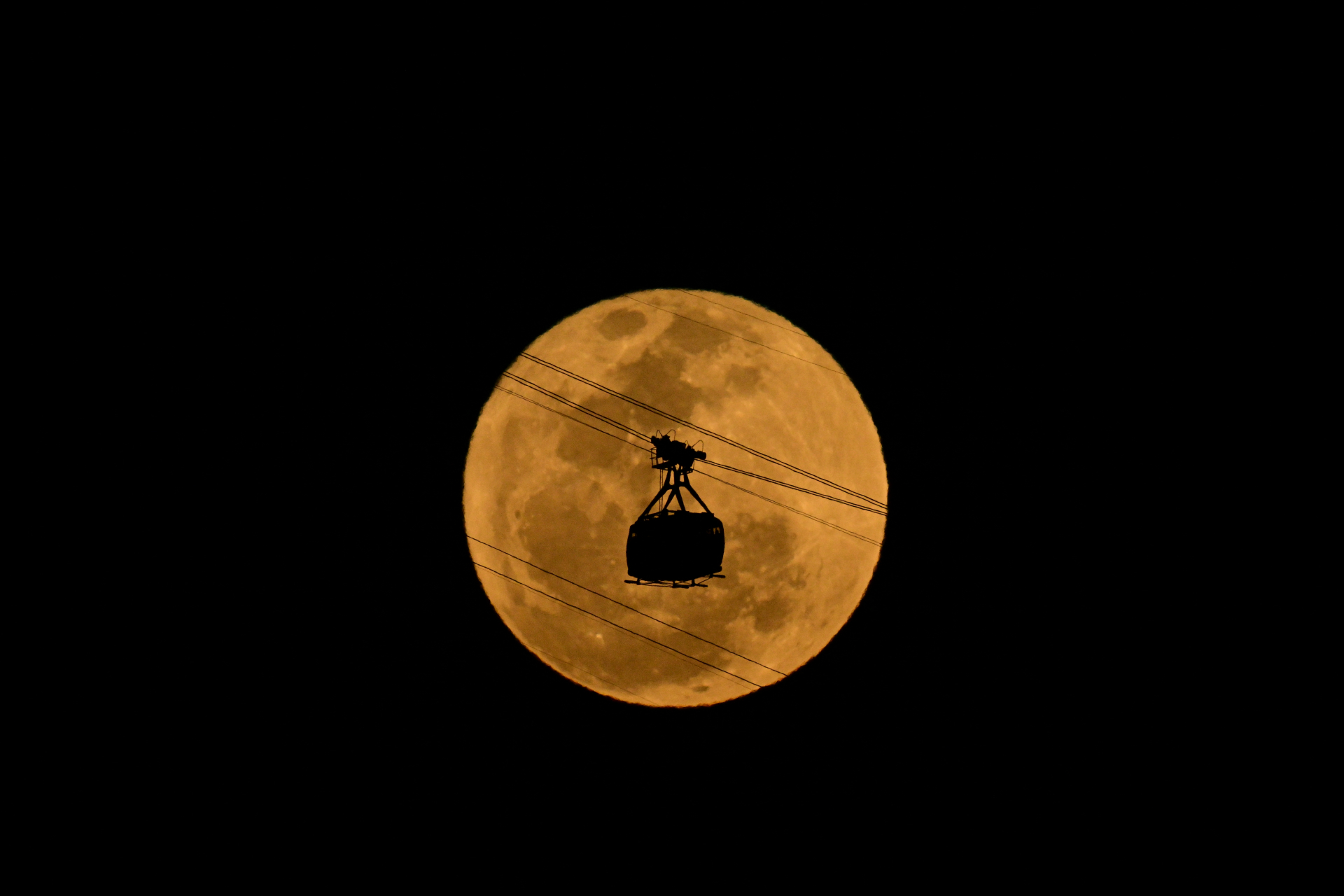 Uma lua cheia grande e brilhante com um leve tom laranja no fundo e a silhueta de um teleférico na frente.