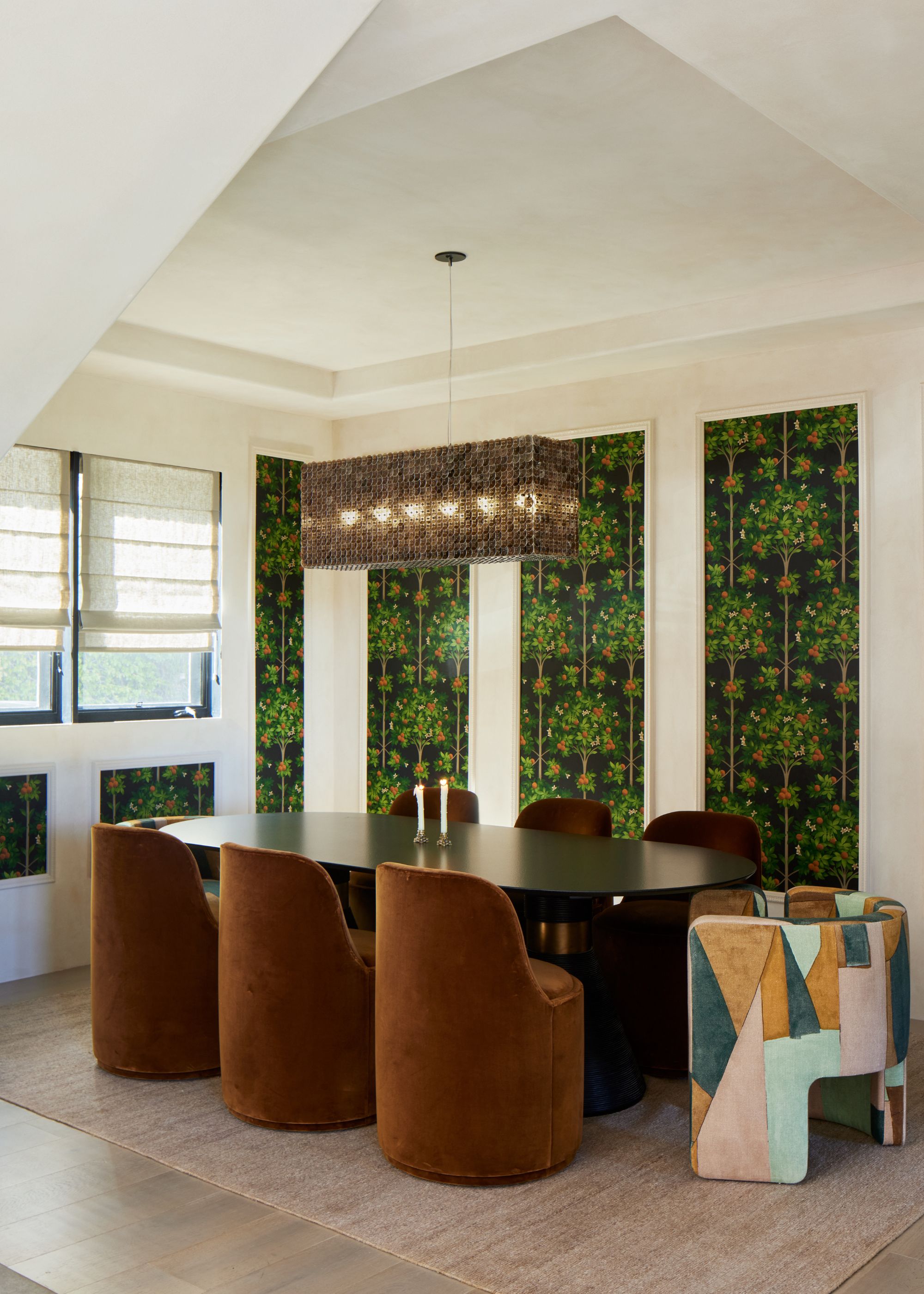 A dining room with white walls and a ceiling in a plaster finish. Four wall panel inserts covered with dark green botanical wallpaper, a large modern dining table, and chocolate brown velvet chairs.