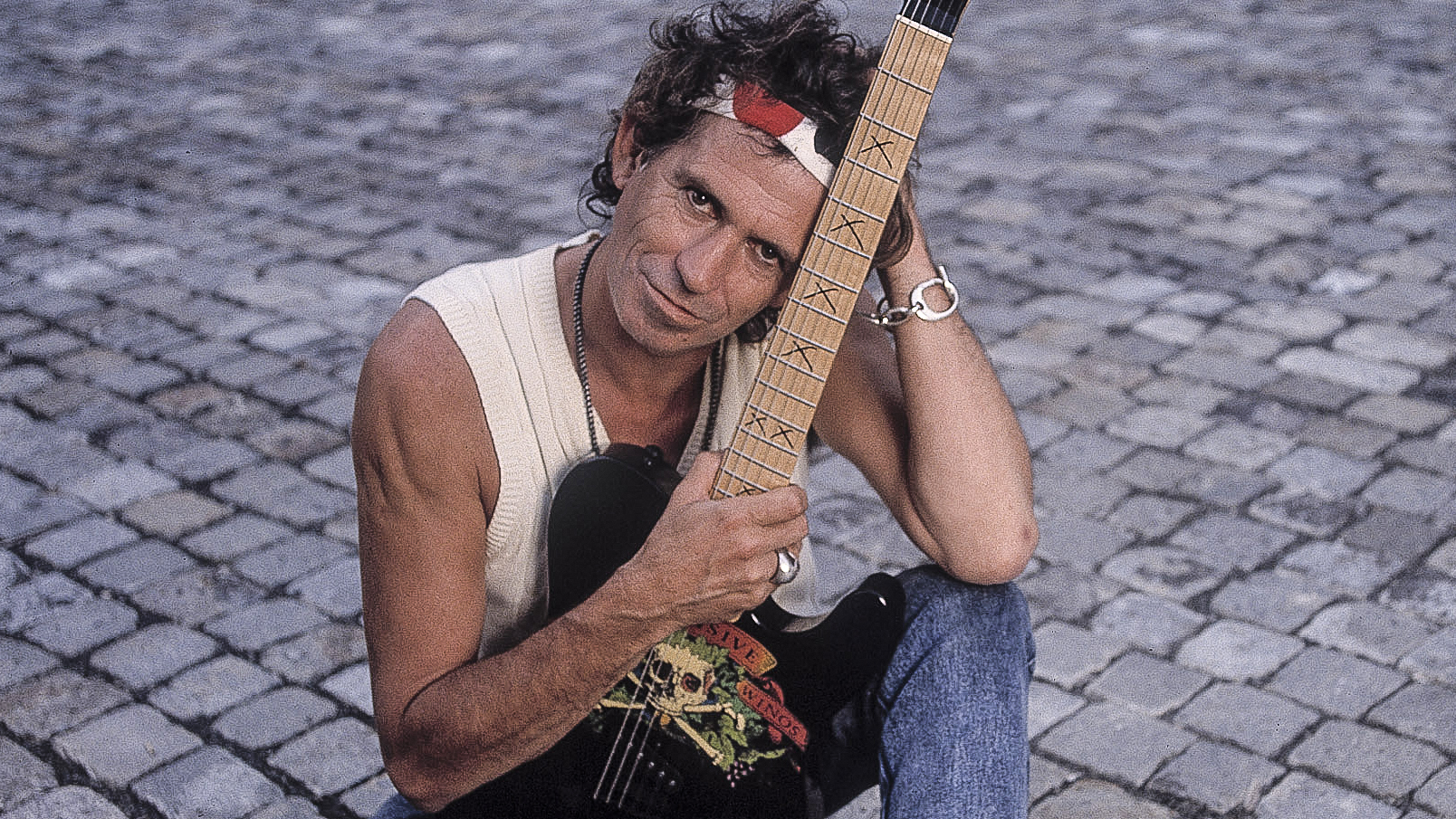 Portrait of British musician Keith Richards as he holds a guitar and sits on a sett-paved driveway, around the time of his 'Talk is Cheap' tour, late 1988.