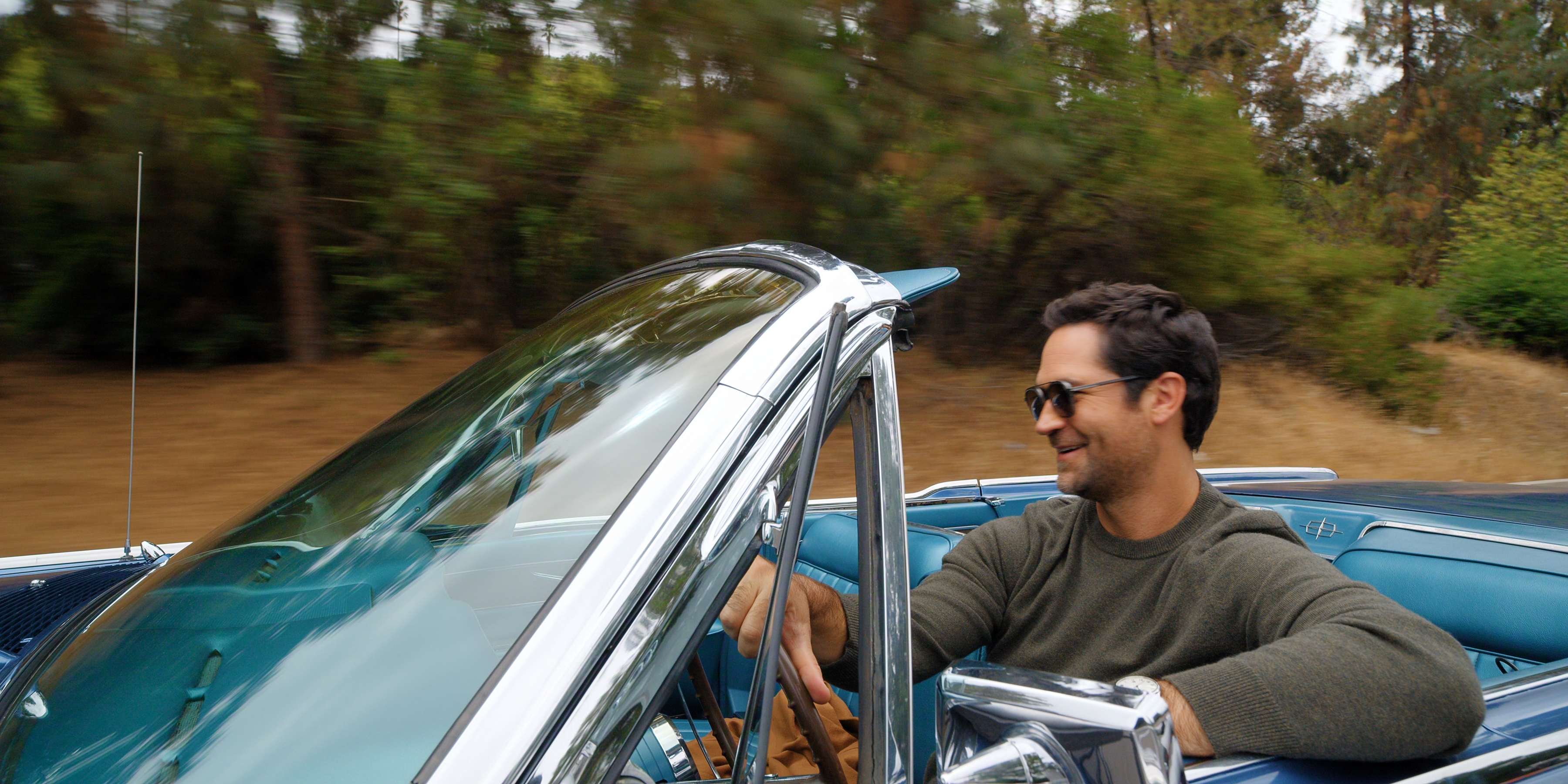 Manuel Garcia-Rulfo as Mickey Haller smiling and wearing sunglasses as he rides in a blue lincoln convertible down a tree-lined road in a still from season 4 of The Lincoln Lawyer