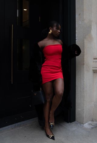 A woman standing in a doorway wearing a black coat over a red minidress and black heels.