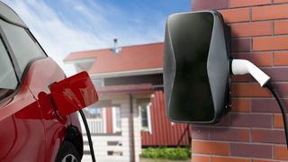 EV charger on a red brick wall plugged into a red car with a house in the background